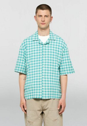 Homme aux cheveux courts portant une chemise à manches courtes à carreaux turquoise avec boutons et un pantalon beige usé, debout devant un fond uni.