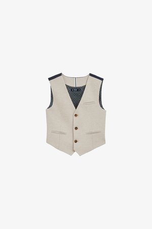 Šviesiai ruda liemenė su V formos iškirpte, trimis rudos spalvos mygtukais ir dviem priekinėmis kišenėmis. Juoda raštuota pamušalo medžiaga matoma viduje, su tamsiomis pečių dalimis.