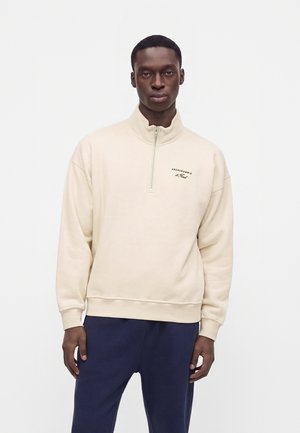 Mann trägt einen beigen Sweatshirt mit Viertel-Reißverschluss und kleinem Logo auf der Brust sowie dunkelblaue Hose und steht vor weißem Hintergrund.