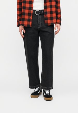 Personne portant un jean noir à jambes larges, une ceinture noire, une chemise à carreaux rouge et noir, un t-shirt blanc en dessous, et des baskets noires avec des lacets blancs.