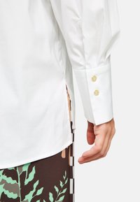 Weißes Langarmshirt mit einem Knopfmanschette mit goldenen Knöpfen und einem seitlichen Schlitz, kontrastierend mit einem gemusterten braunen Unterteil.