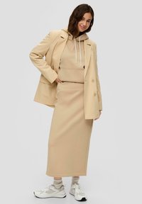 Beige hoodie met trekkoorden onder een licht gestreepte blazer, gecombineerd met een bijpassende rok en witte sneakers. Glad, zacht materiaal.