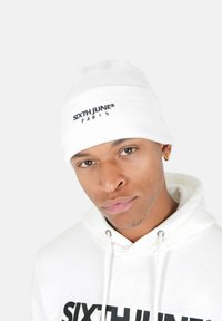 Vit stickad mössa med nederkant, med svart broderi av "SIXTH JUNE® PARIS". Materialet ser mjukt och texturerat ut.