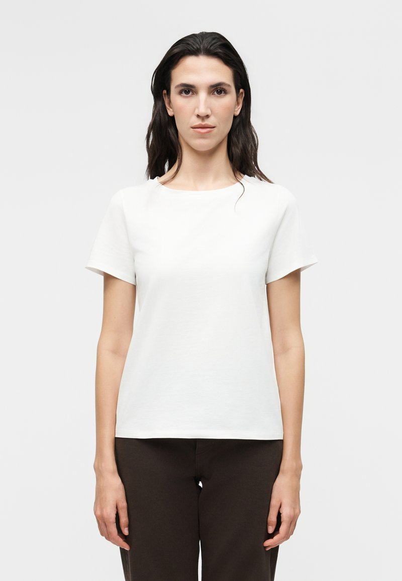 Femme aux longs cheveux foncés portant un t-shirt blanc uni à manches courtes et un pantalon foncé, debout devant un fond gris clair.