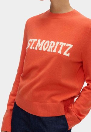 Person iført en orange langærmet sweater med teksten "ST. MORITZ" i hvidt hen over brystet og mørkeblå bukser.