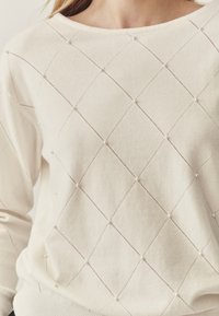 Világos bézs pulóver gyémánt kötésmintával, apró gyöngyszerű részletekkel díszítve és kerek nyakkivágással. Puha textúrájú és laza szabású.