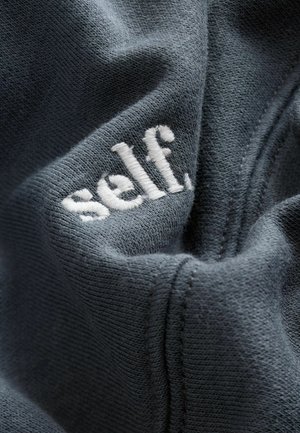 Tamno sivi materijal ima teksturiranu površinu s istaknutim bijelim izvezenim logom koji glasi "self." Vidljive šavove ocrtavaju dizajn.