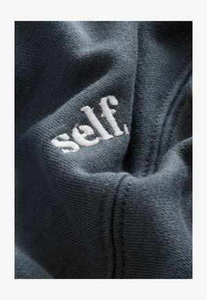 Donkergrijze stof heeft een gestructureerde oppervlakte met een opvallend wit geborduurd logo dat "self" leest. Zichtbare stiksels omlijnen het ontwerp.