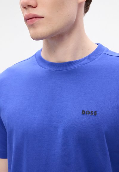 T-shirt en coton bleu avec un col rond et des manches courtes. Présente un logo noir "BOSS" sur la partie gauche de la poitrine. Texture lisse.