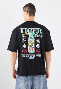 T-shirt noir avec un design graphique coloré au dos, représentant une bouteille de soda, un tigre et du texte en japonais. Coupe ample, manches courtes.