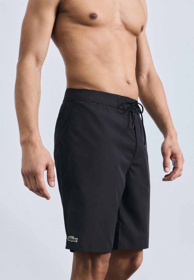 Γυμνός άνδρας που φοράει μαύρο μαγιό με κορδόνι και πράσινο λογότυπο κροκοδείλου στο αριστερό πόδι, σε ουδέτερο φόντο.