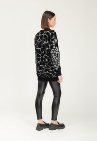 Donna in un maglione a motivo giraffa nero e grigio, leggings neri in ecopelle e mocassini neri chunky, in piedi di lato su uno sfondo neutro.