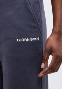 Prim-plan cu pantaloni albastru închis cu logo alb „BJÖRN BORG” lângă buzunar, o mână de culoare închisă se odihnește lângă pantaloni.