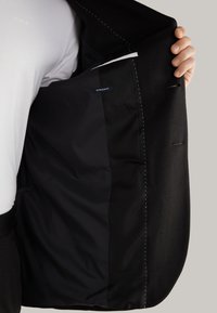 Schwarzer Blazer mit glatter Textur, der ein weißes Innenfutter, ein Etikett für besonders schlanke Passform und kontrastierende Nähte entlang der Kanten aufweist.