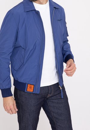 Mand iført en blå bomberjakke over en hvid t-shirt og mørke jeans, med lynlåsen delvist åben og hænderne nær taljen.