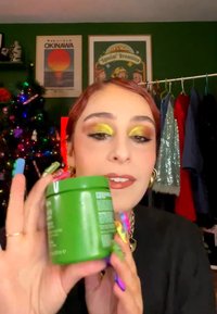 Femme avec un fard à paupières jaune scintillant tient un pot vert, avec des ongles colorés, un sapin de Noël, des affiches et un portant à vêtements dans une pièce verte derrière elle.