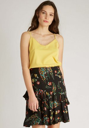 Top en satin jaune avec des empiècements en dentelle, associé à une jupe à volants noire à motifs floraux, ornée de fleurs colorées et de feuillage vert.