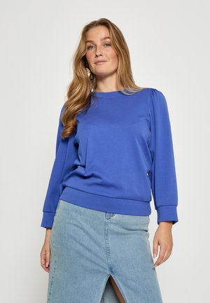 Felpa blu con maniche lunghe, polsini e orlo a costine. Indossata con una gonna di jeans, caratterizzata da uno spacco frontale e una tonalità chiara.