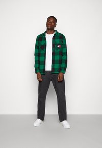 Camicia di flanella a quadri verdi e neri con due tasche sul petto, indossata sopra una maglietta bianca. Abbinata a jeans grigio scuro e sneaker bianche.