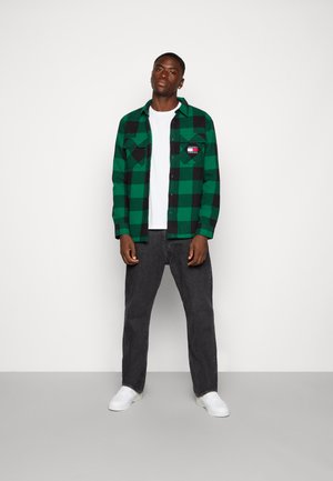 Camicia di flanella a quadri verdi e neri con due tasche sul petto, indossata sopra una maglietta bianca. Abbinata a jeans grigio scuro e sneaker bianche.