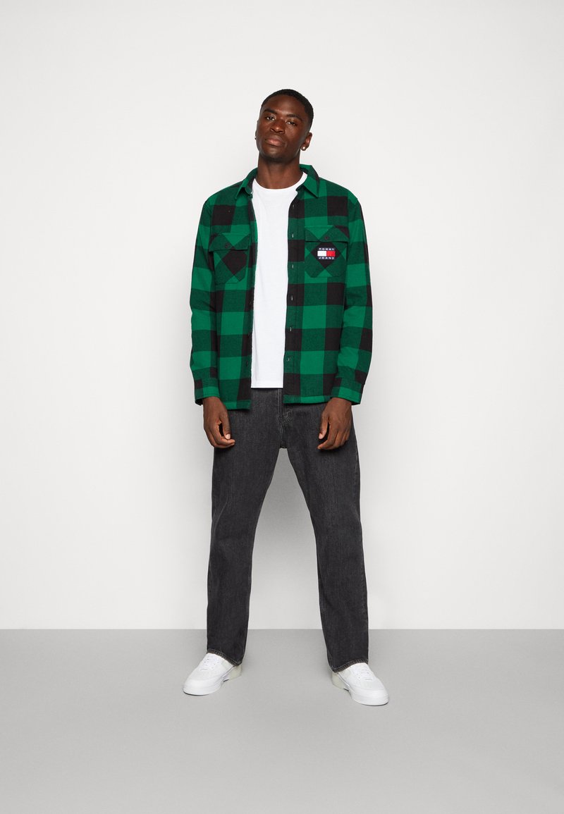 Camicia di flanella a quadri verdi e neri con due tasche sul petto, indossata sopra una maglietta bianca. Abbinata a jeans grigio scuro e sneaker bianche.