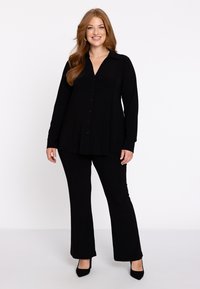 Schwarze Bluse mit langen Ärmeln, Knopfleiste und spitzem Kragen, kombiniert mit schwarzen Schlaghosen und schwarzen High Heels. Glatte Stoffstruktur.