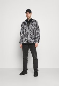 Lichte jas in een zwart-wit abstract patroon, met een rits aan de voorkant, verstelbare capuchon en elastische manchetten, gecombineerd met zwarte jeans en sneakers.