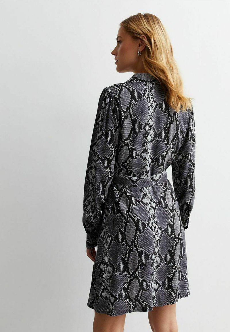 Schwarz-weißer Schlangenprint-Kleid mit langen Ärmeln, knöpfbarem Kragen und einem passenden Gürtel in der Taille. Der Stoff wirkt glatt mit einem leichten Glanz.