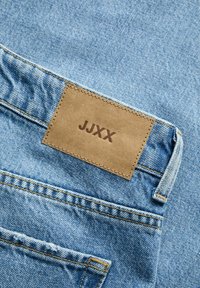 Μπλε τζιν denim σε κοντινό πλάνο που δείχνει την πίσω τσέπη και καφέ δερμάτινη ετικέτα με κεντημένο το κείμενο "JJXX" πάνω από αυτή.