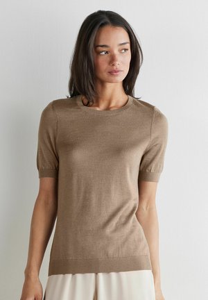 Frau mit glatten, dunklen Haaren, die ein eng anliegendes, hellbraunes kurzärmeliges Strickoberteil und einen cremefarbenen Rock trägt, steht vor einer schlichten Wand.