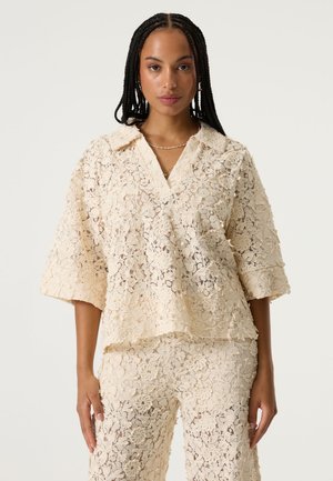 Spitzebluse in Cremefarbe mit lockerem Schnitt, weiten Ärmeln und einem Kragen, versehen mit Blumenmustern und transparenter Textur.