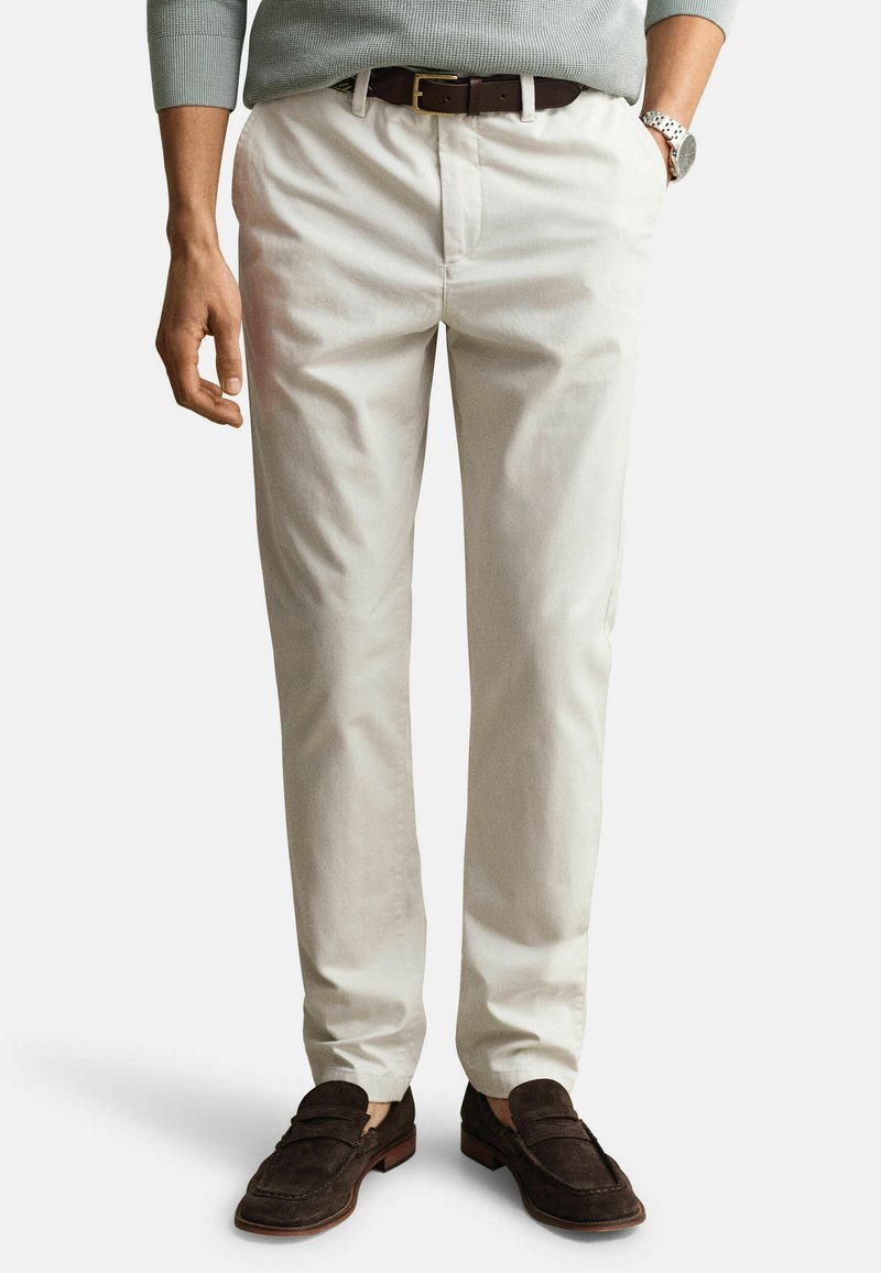 Homme portant un pantalon chino beige, une ceinture marron foncé, un pull gris, des mocassins marron foncé et une montre-bracelet argentée, debout avec les mains détendues.