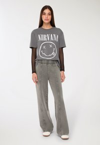 Młoda kobieta ubrana w szary t-shirt Nirvana na czarną bluzkę z długim rękawem, luźne szare spodnie i białe buty, stojąca na białym tle.