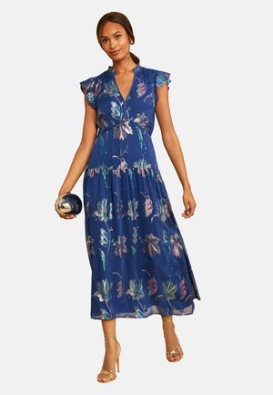 Donna che indossa un vestito midi blu a fiori con maniche a volant, tiene in mano una pochette tonda metallica e indossa tacchi alti dorati con cinturini.