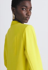 Bright yellow blazer with structured shoulders, smooth fabric, and tailored fit. Model features gold accented jewelry and short hair.