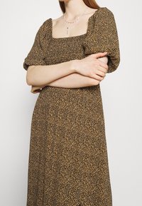 Ruskea, lyhythihainen mekko, jossa on neliön muotoinen pääntie ja keltaisia sekä valkoisia kukkakuoseja. Mekossa on löysä siluetti ja teksturoitu kangas.