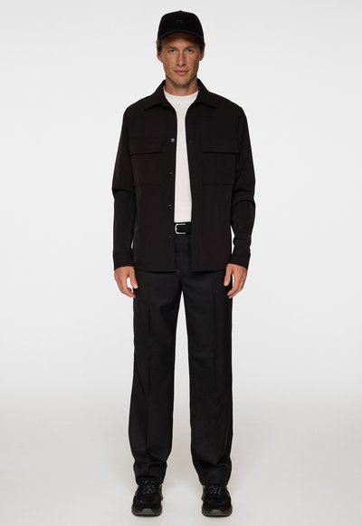 Svart skjorta med knappar och två bröstfickor ovanpå en vit T-shirt, i kombination med svarta byxor och svarta sneakers, mot en vit bakgrund.