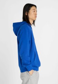 Felpa blu realizzata in tessuto morbido; presenta una tasca a marsupio e un cappuccio con cordini. Indossata con pantaloni sportivi grigi, esposta su uno sfondo bianco.