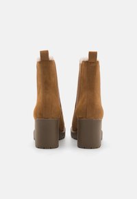 Botas de ante marrón hasta el tobillo con un tacón apilado y un interior suave. Cuenta con una lengüeta en la parte trasera y costuras laterales suaves.