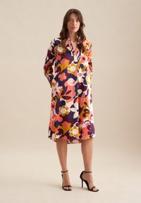 Langes Hemdblusenkleid mit floralen Mustern in Rosa, Rot, Gelb und Weiß. Hergestellt aus leichtem Stoff, mit Kragen und langen Ärmeln.