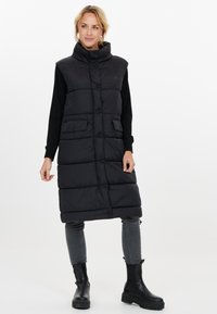 Lång svart vadderad väst med hög krage, knappstängning, två framfickor och rak siluett, bärs över en svart topp och grå jeans.