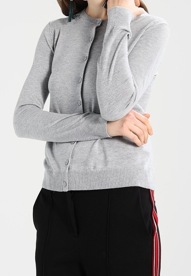 Kvinna iklädd en ljusgrå, knäppt cardigan och svarta byxor med röda och vita sidogram. Hon poserar med armarna i kors.