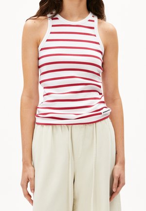 Femme portant un haut blanc sans manches avec des rayures horizontales rouges et un pantalon beige ample, debout avec les bras détendus sur les côtés.