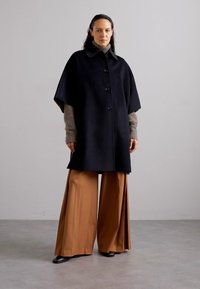 Cappotto in lana nera con colletto rotondo e bottoni frontali, abbinato a pantaloni marroni a gamba larga e maniche grigie a coste. Texture liscia.