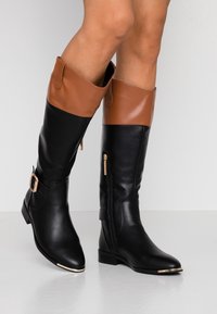 Piernas con botas de cuero altas negras y marrones, con cremalleras laterales, hebilla dorada y acentos metálicos en la punta, sobre un suelo claro.