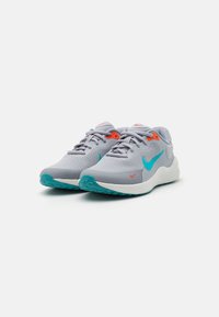 Zapatillas deportivas gris claro con acentos en azul verdoso y detalles en naranja. Presentan una parte superior de malla transpirable y una suela de goma texturizada. Logo de Nike visible.