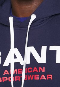 Sudadera con capucha de la Marina hecha de mezcla de algodón con cordones blancos. Presenta un texto grande impreso "GANT" en blanco y "AMERICAN SPORTSWEAR" en rojo.