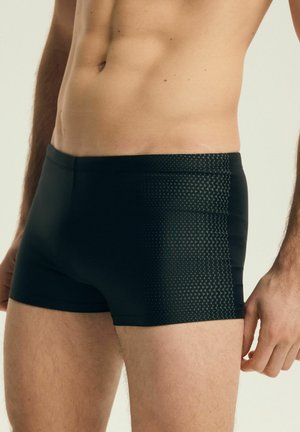 Pánský trup v černých plavkách s jemným trojúhelníkovým vzorem na pravé straně, stojící na světlém pozadí.