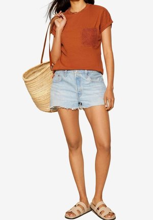 Femme portant un t-shirt orange rouille avec une poche en dentelle, un short en jean clair, des sandales beiges et portant un grand sac cabas en paille tressée sur l'épaule.