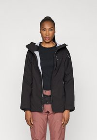 Jachetă neagră impermeabilă cu glugă, căptușeală gri, fermoar frontal și multiple buzunare cu fermoar. Pur tată pe un top negru și pantaloni roz.